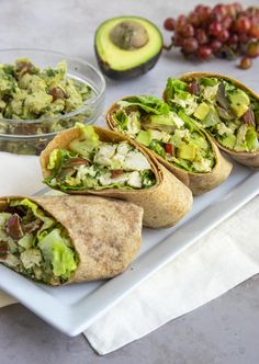 there are four burritos on the plate with grapes and avocado in the background