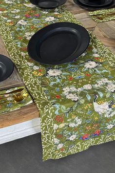 the table is set with black plates and placemats on green floral patterned cloth