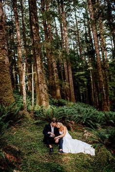 a bride and groom sitting on the ground in front of some tall trees at their wedding