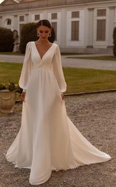 a woman standing in front of a large house wearing a white wedding dress with long sleeves