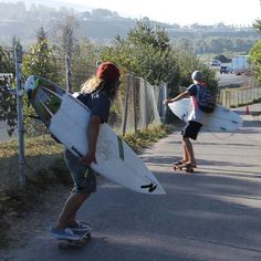 two people with surfboards walking down the side of a road in front of a fence