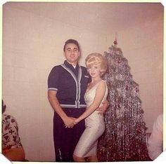 a man and woman standing next to a christmas tree in front of a white wall