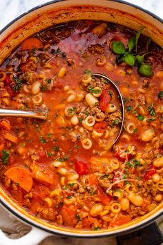 a pot filled with pasta and meat soup on top of a white countertop next to a wooden spoon