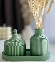 two green vases sitting on top of a white table next to each other with dried grass in them