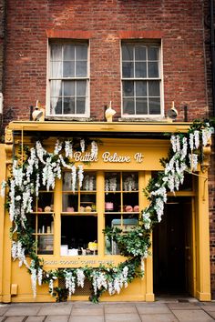 a yellow store front with white flowers and greenery on the outside, in front of a brick building