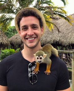 a man with a hat and sunglasses holding a small monkey on his shoulder in front of palm trees