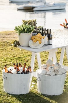 two white buckets filled with wine bottles next to an outdoor table full of food and drinks