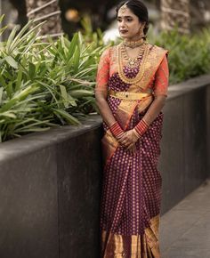 a woman in a purple and gold sari standing next to a planter with greenery