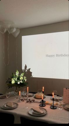 a table set for a birthday party with candles and flowers on it, in front of a projector screen