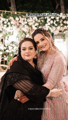 two women hugging each other in front of a flower covered area with flowers on the wall behind them