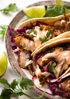 three fish tacos on a plate with limes and cilantro