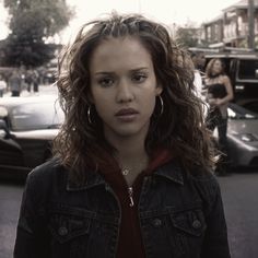 a woman standing in the middle of a street with cars behind her and people walking on the sidewalk