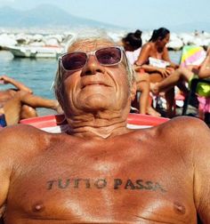 an older man with tattoos on his chest sitting in front of the water and people