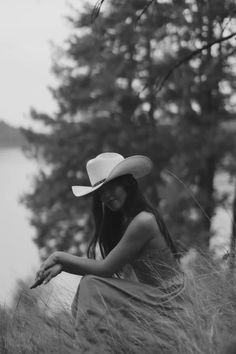 a woman wearing a cowboy hat sitting in tall grass by the water's edge