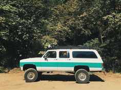 a white and blue truck parked in front of some trees on the side of a dirt road