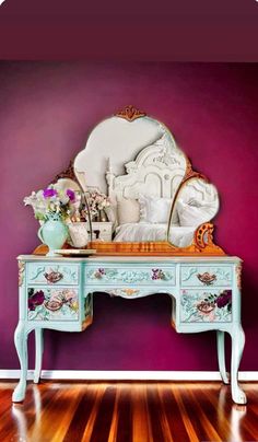 an old dresser with a mirror and flowers on it in front of a purple wall