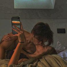 a man and woman laying in bed kissing while holding up a cell phone to their face