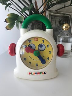 a white clock with a green handle sitting on top of a table next to a potted plant