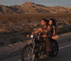 a man and woman riding on the back of a motorcycle down a road in the desert