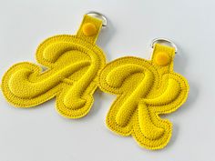 two yellow crocheted keychains with letters on the front and one in the back