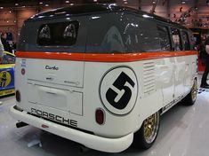 an orange and white vw bus on display in a building with people looking at it