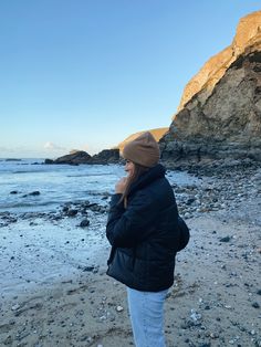 a woman standing on top of a sandy beach next to the ocean wearing a black jacket