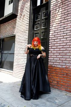 a woman with red hair in a black dress standing on the sidewalk next to a brick building