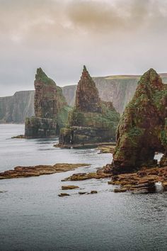 some very big rocks in the water by some cliffs and grass on the shore with an overcast sky