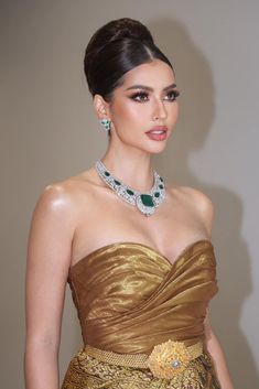 a woman wearing a gold dress and necklace with jewels on her neck, standing in front of a wall