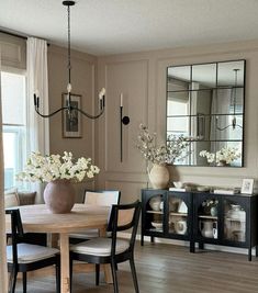 a dining room table and chairs with flowers in vases on the sideboard next to it