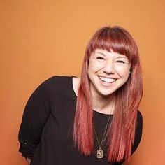 a woman with long red hair smiling at the camera while standing in front of an orange wall