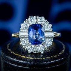 A stunning Art Deco-inspired sapphire diamond cluster ring adorned with a deep-blue cushion cut sapphire in the centre, framed by eight round transitional cut diamonds and two baguette cuts. The sapphire weighs approx. 2ct and is complemented beautifully by the bright, sparkling diamonds that have SI1 clarity – H colour, approx. 1ct total. The sapphire is claw set in a four-prong mount atop a platinum face with an 18ct yellow gold back and band. It’s a magnificent Art Deco-style statement pi... Edwardian Engagement Ring, Uk Products, Sapphire Diamond Ring, Diamond Cluster Ring, Art Deco Inspired, Memento Mori, Style Statement, Diamond Cluster