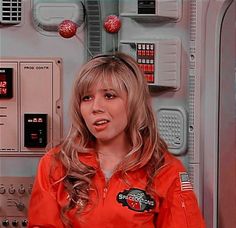 a woman in an orange space suit standing next to a control panel with red balls on it