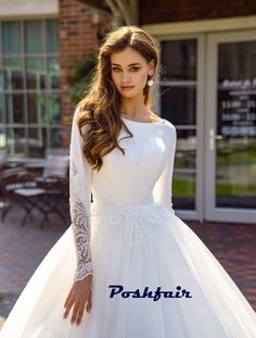 a woman wearing a white wedding dress with long sleeves and lace on the top, standing in front of a brick building