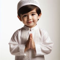 a little boy wearing a white outfit and standing in front of a white background with his hands together