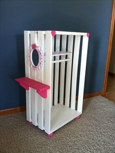 a white wooden toy cage with pink trim on the bottom shelf and mirror mounted to it's side