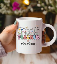 a person holding a white coffee mug with the words teacher on it and colorful flowers