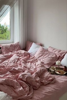 an unmade bed with pink sheets and pillows in front of a large open window