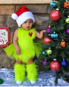 a baby dressed up in a christmas outfit standing next to a tree with decorations on it