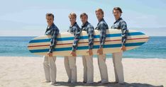 four men in plaid shirts are holding a surfboard on the beach while standing next to each other