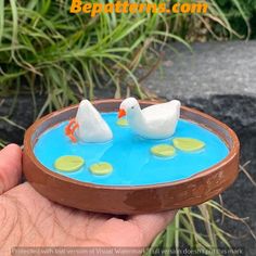 a hand holding a miniature model of two white ducks in a blue pond with green leaves