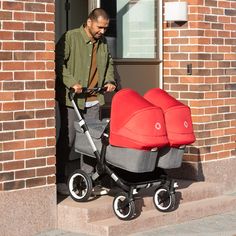 a man pushing a stroller next to a red and gray baby buggy in front of a brick building