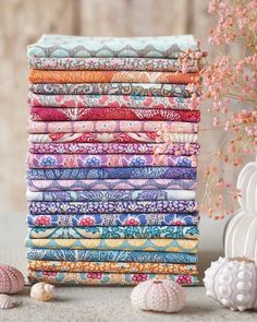 a stack of colorful fabrics next to seashells and shells on a table with flowers in the background