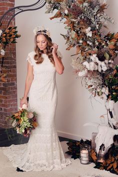 a woman standing in front of a floral arch wearing a white dress and holding flowers