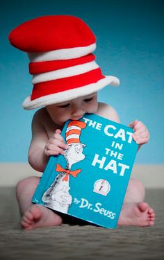 a baby wearing a red and white cat in the hat holding a blue book with dr seuss written on it