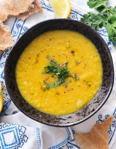 a black bowl filled with yellow soup next to some bread and cilantro leaves