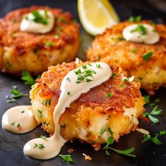 three crab cakes with sauce and garnish on a black plate next to lemon wedges