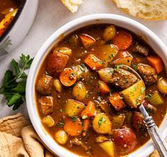 a white bowl filled with stew next to bread and parsley on a table top