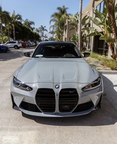 the front end of a silver car parked in a parking lot next to palm trees