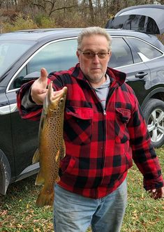 a man holding a fish in front of a parked car and giving the thumbs up sign